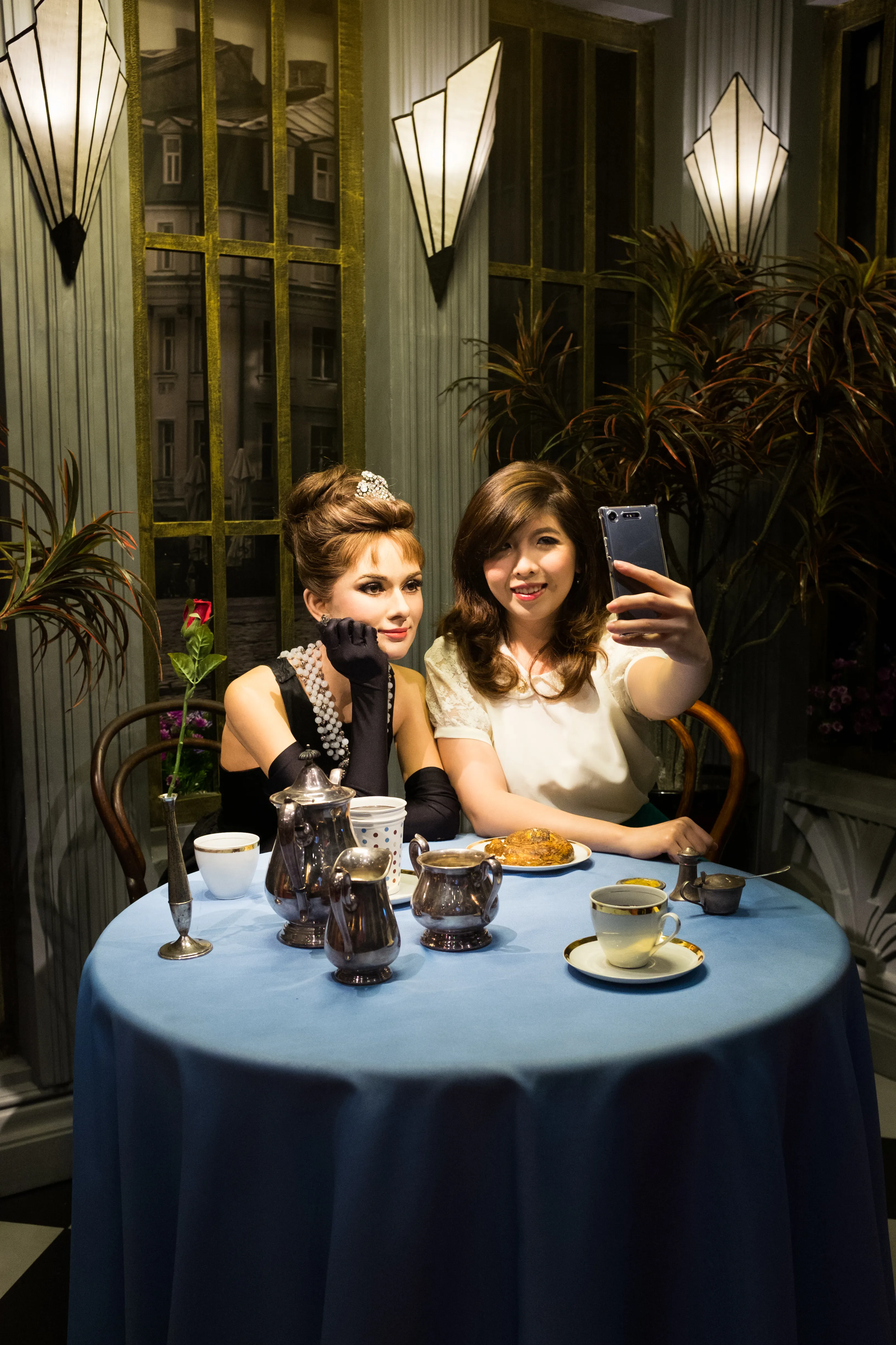 A woman taking a selfie with a wax figurine of Audrey Hepburn at Madame Tussauds Singapore TV & Film Zone. The wax figurine is dressed in a black gown with pearl earrings and necklace. 
